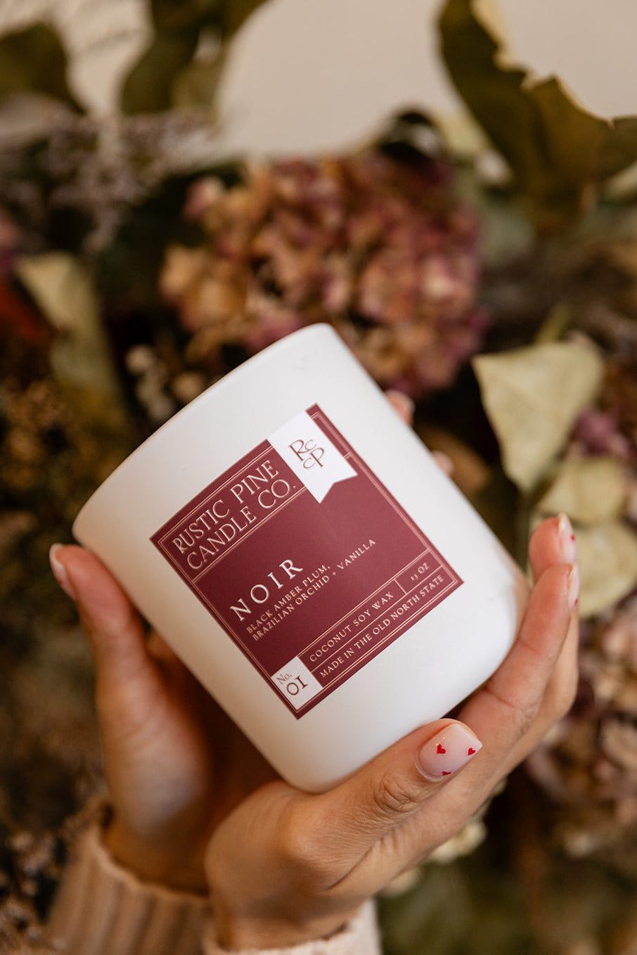 Hand holding a white candle labeled 'Noir' with a blurred floral background